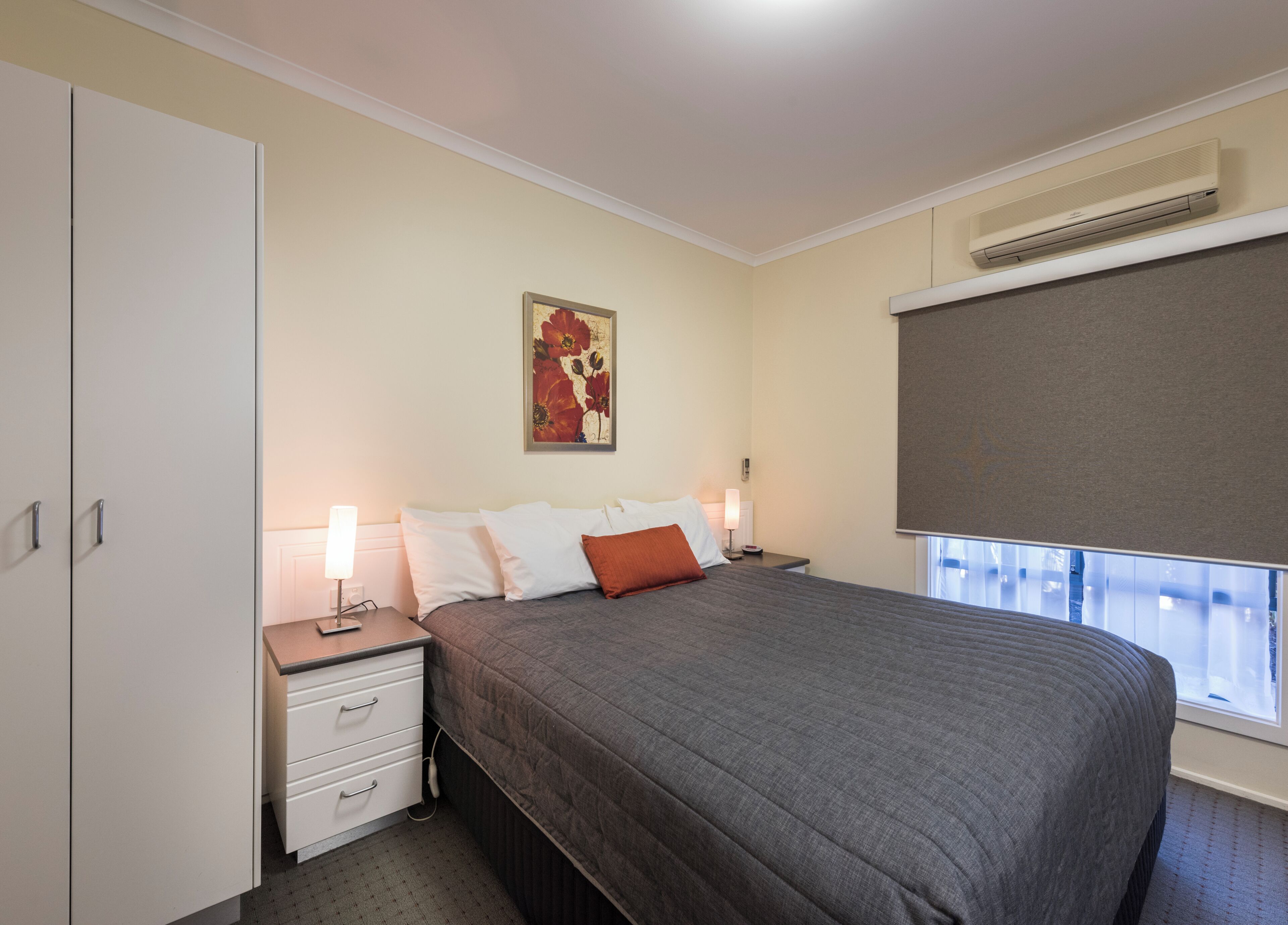 Un dormitorio con cama, mesitas de noche, aire acondicionado instalado en la pared y una ventana con persianas.
