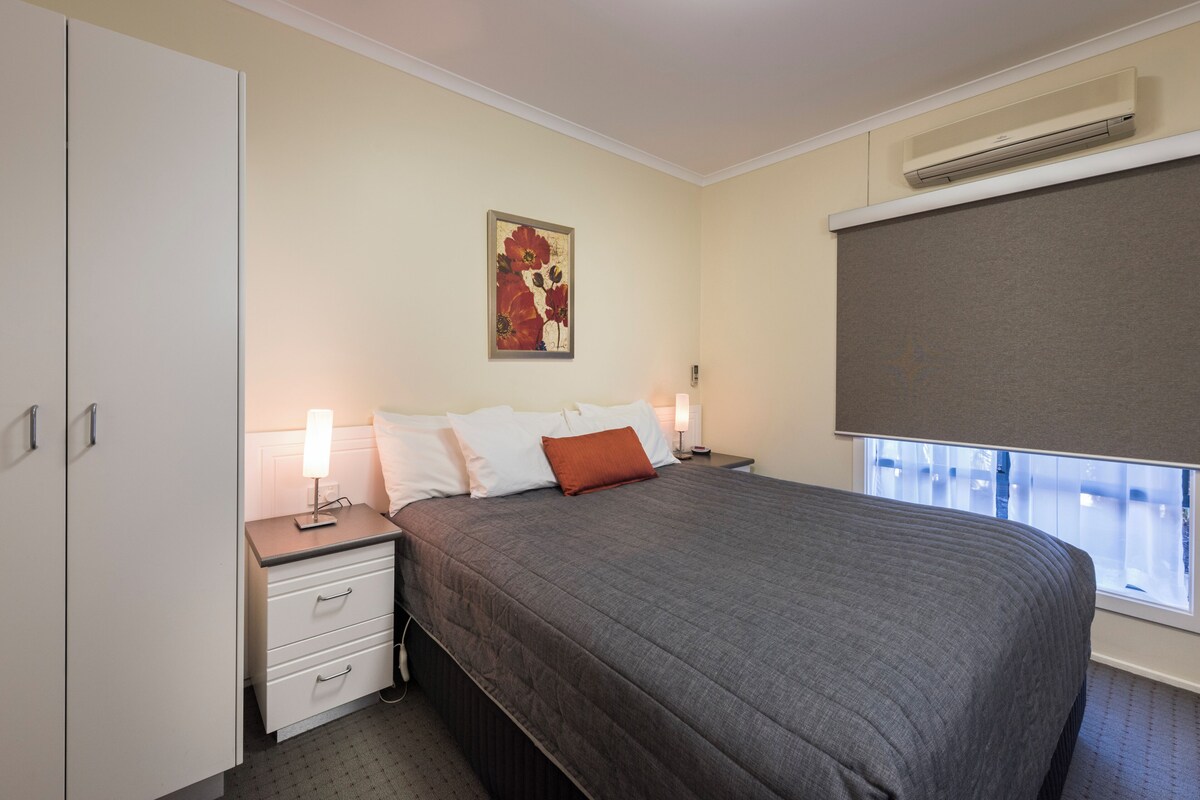 A bedroom with a bed, bedside tables, a nightstand, a wall-mounted air conditioner, and a window with blinds.