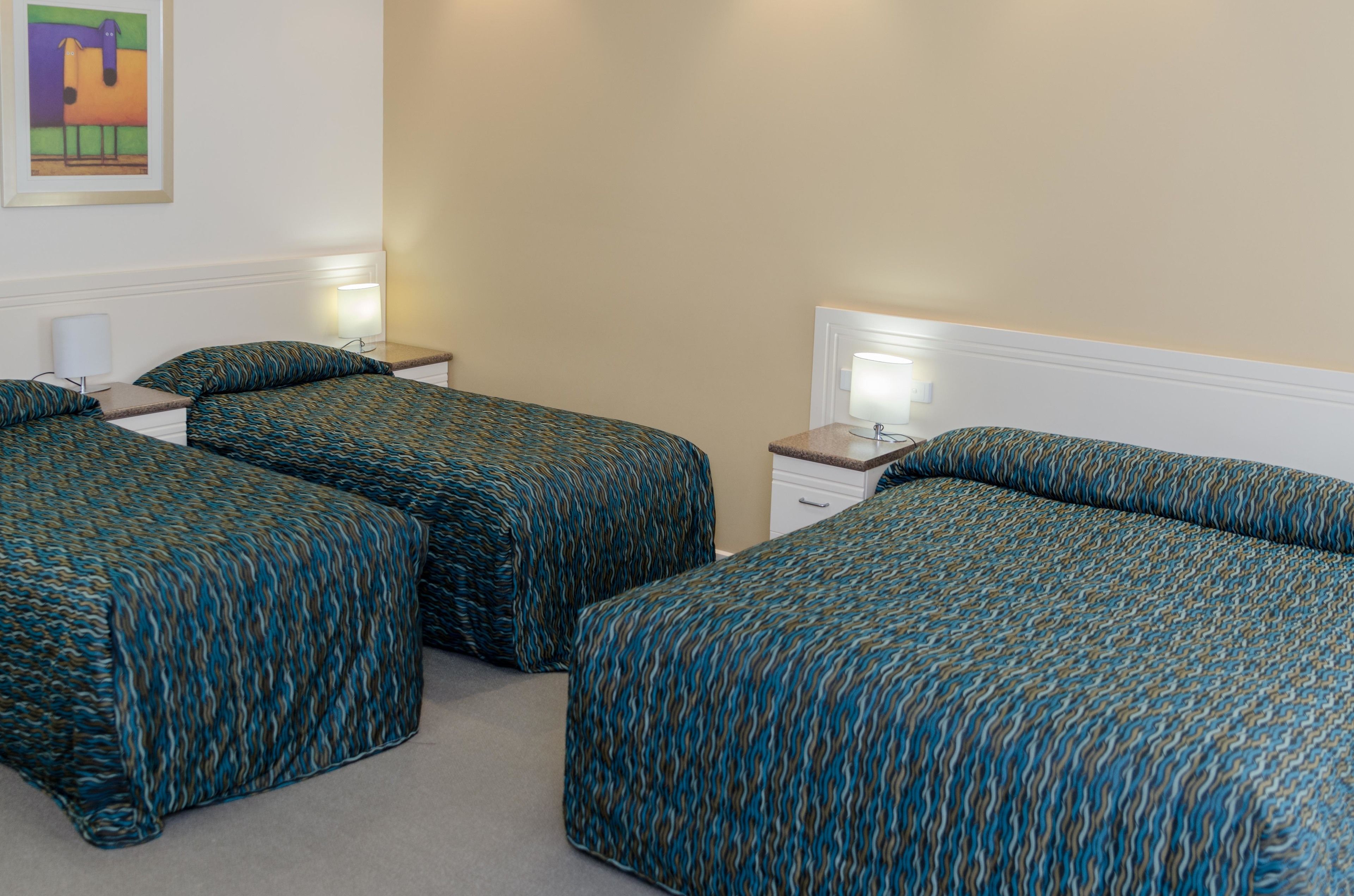 A hotel room with three beds, each with a patterned bedspread, a bedside table with a lamp, and a wall-mounted artwork.