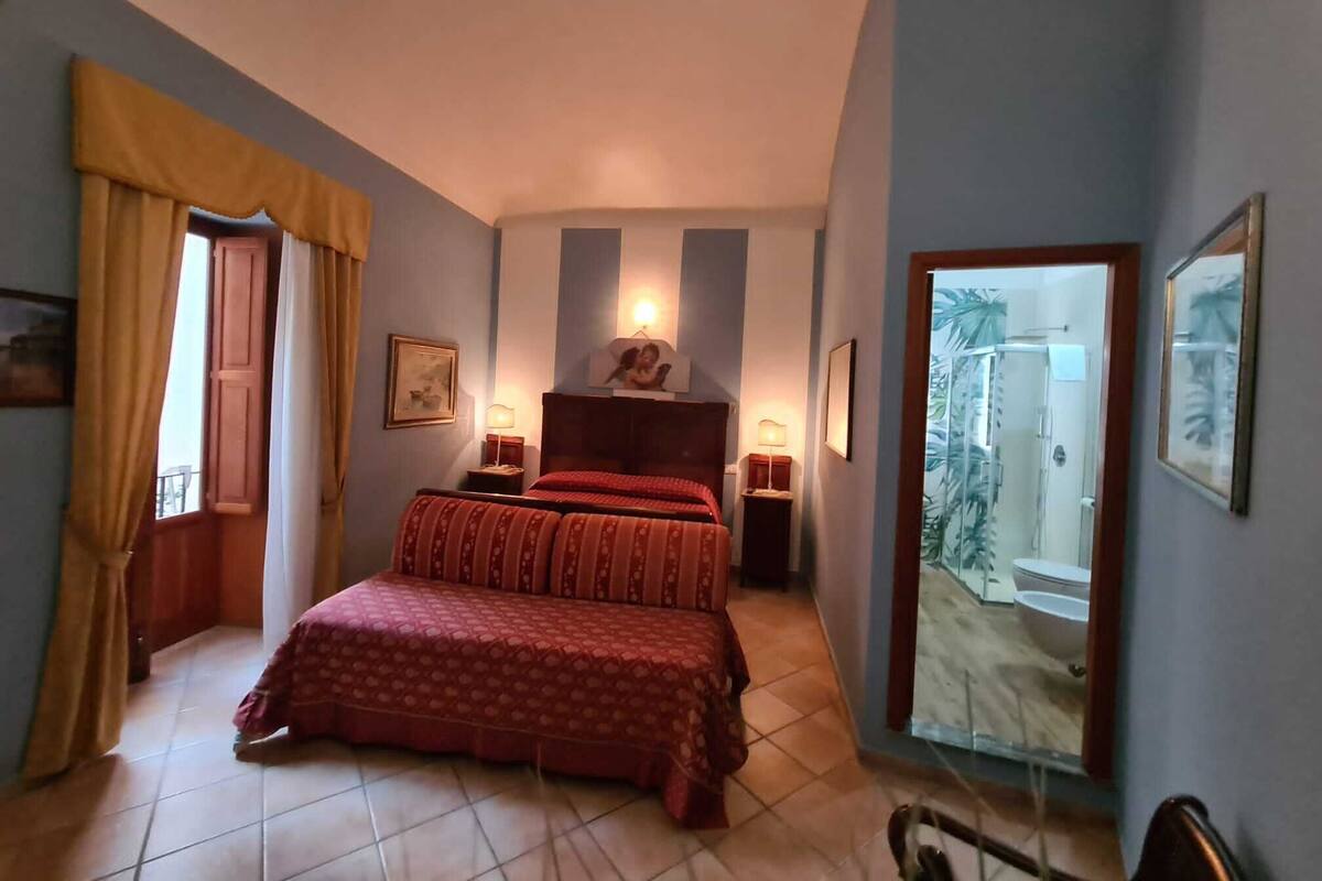 A bedroom with a bed, two bedside tables, a nightstand, a window with curtains, and a door leading to a bathroom.