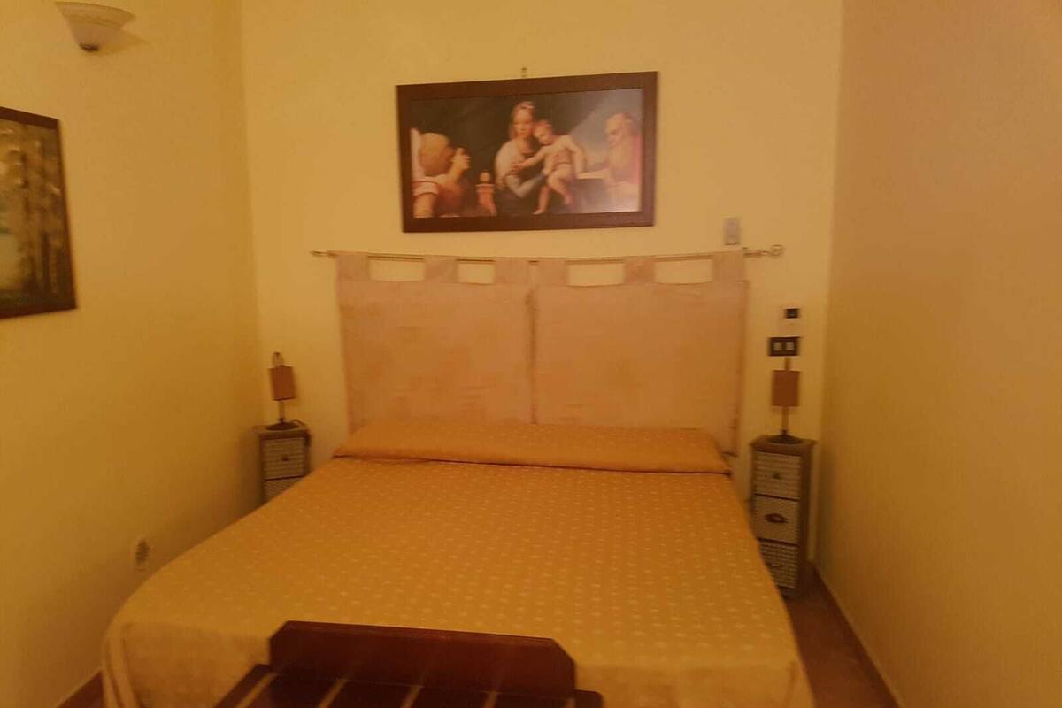 A small, narrow room with a bed, a nightstand, and a wall-mounted lamp.