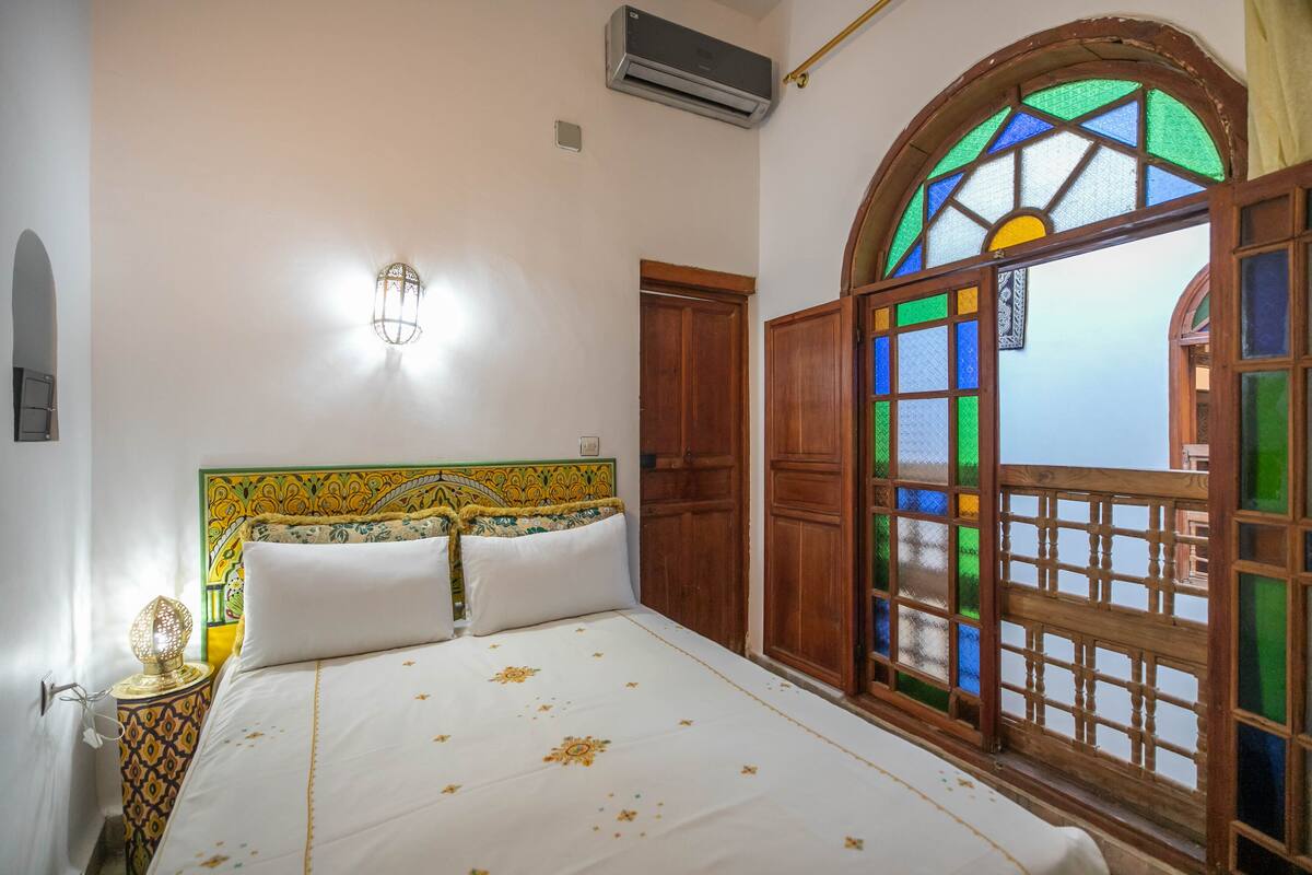 A bedroom with a bed, a nightstand, a wall-mounted lamp, a wooden wardrobe, and a large window with colorful stained glass.