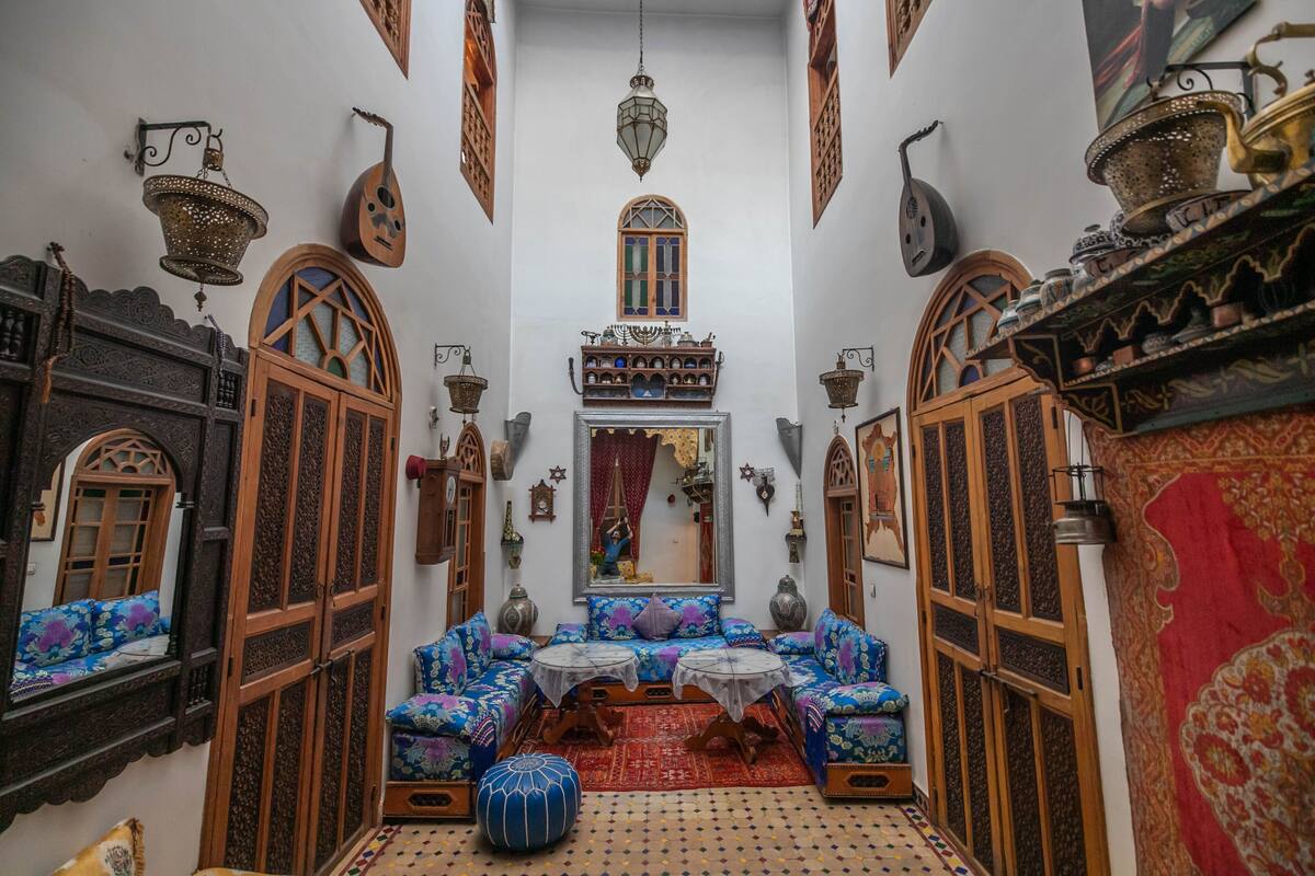 A narrow, ornately decorated hallway with traditional Moroccan architecture, including wooden doors, intricate wall designs, and hanging lanterns.