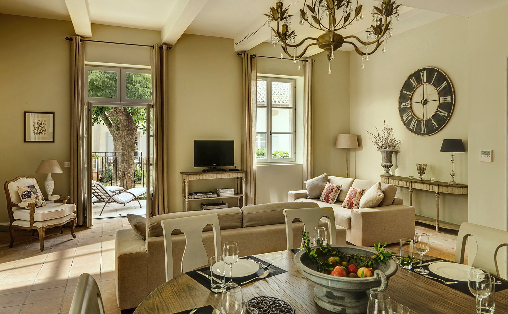 A well-lit living room with a sofa, dining table set for two, a television, and a large wall clock.