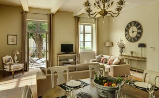 Un salón luminoso con sofá, una mesa de comedor preparada para dos personas, un televisor y un reloj de pared grande.