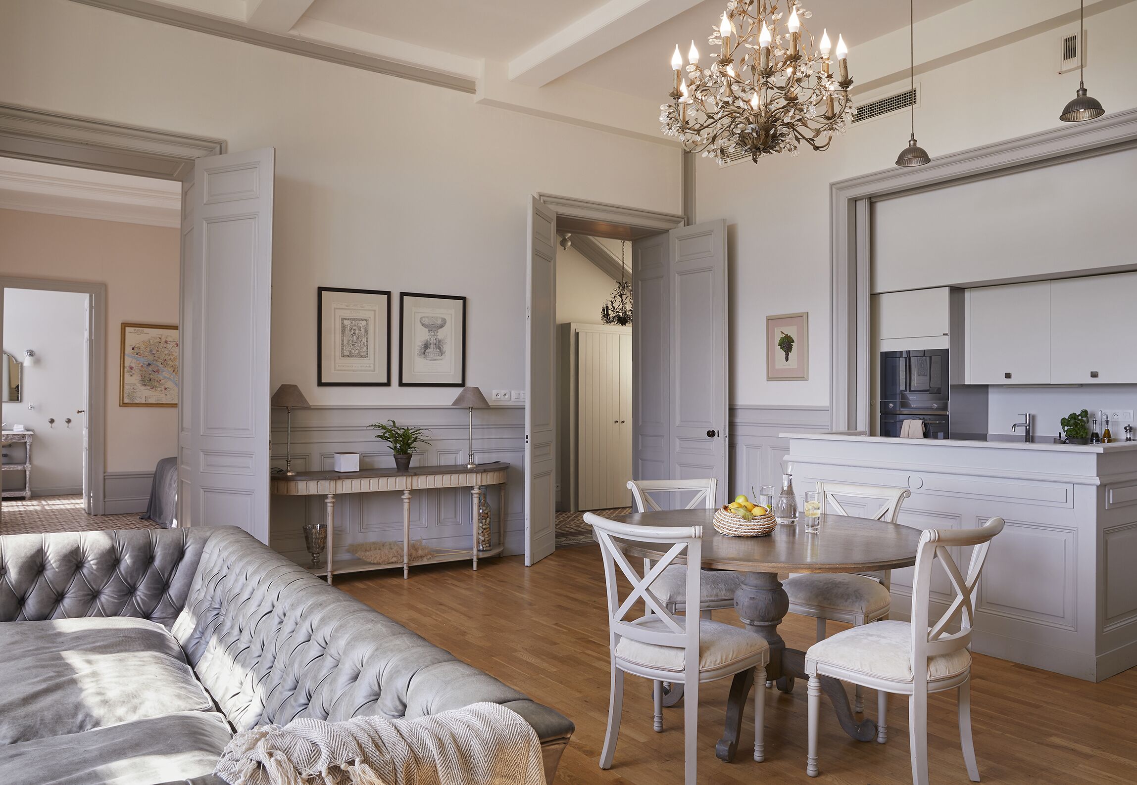 A spacious living room with a grey tufted sofa, a dining area with white chairs, a chandelier, and a kitchen with white cabinetry.