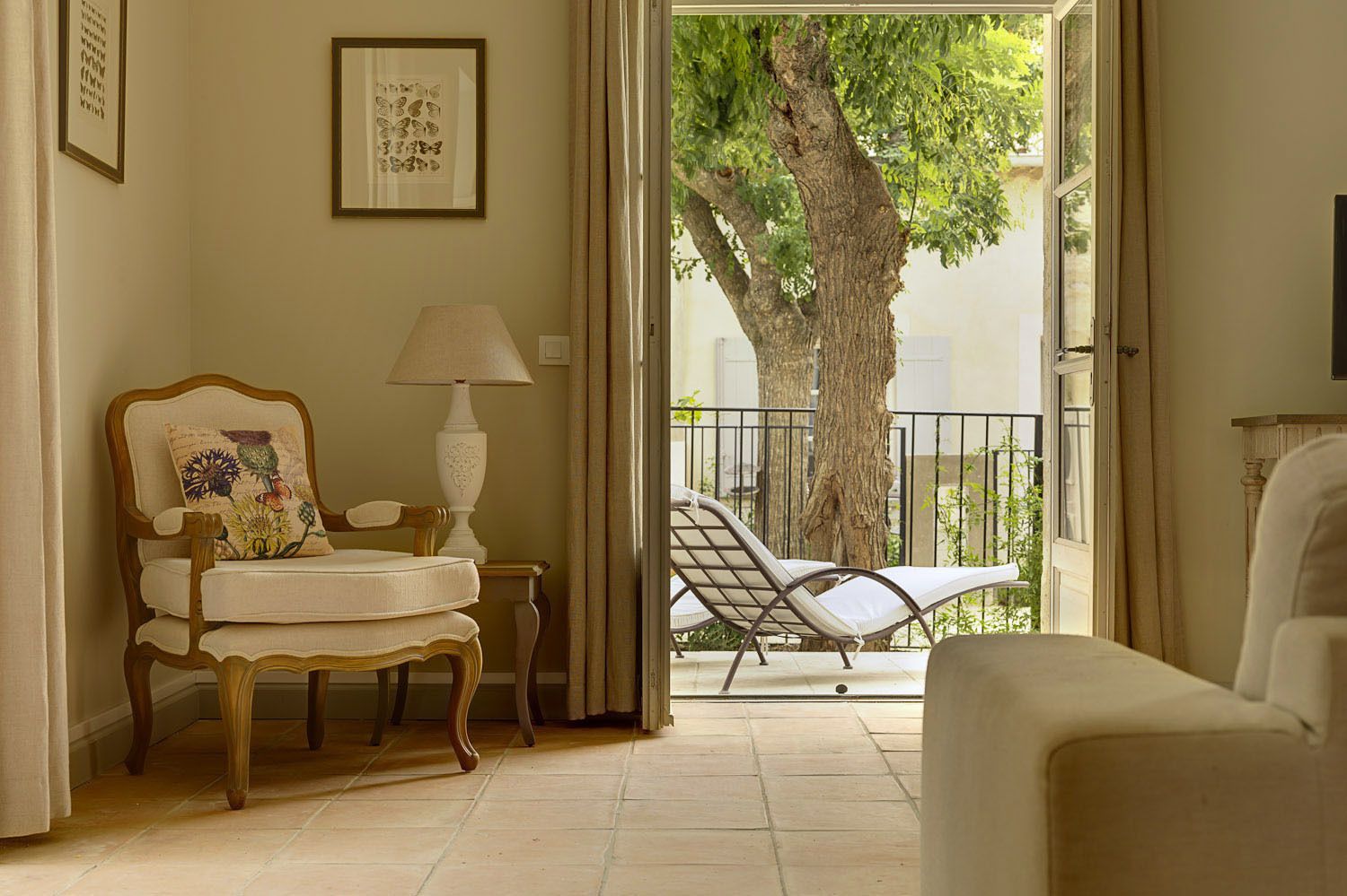 A room with a wooden chair, a table with a lamp, and a lounge chair by a sliding glass door leading to a balcony with a tree.