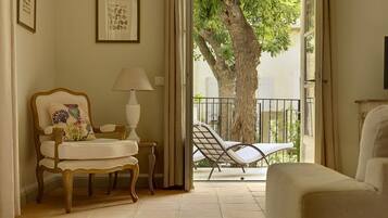 A room with a wooden chair, a table with a lamp, and a lounge chair by a sliding glass door leading to a balcony with a tree.