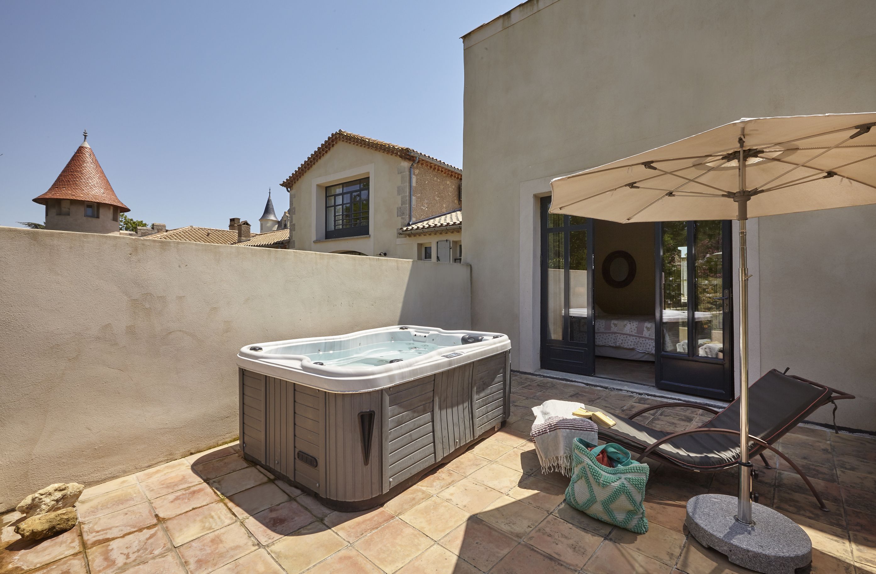 A hot tub in a patio area with a chair and umbrella, a stone wall, and a building with a tiled roof in the background.