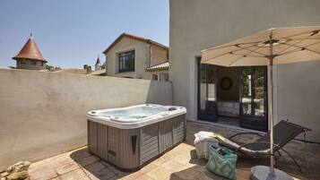 A hot tub in a patio area with a chair and umbrella, a stone wall, and a building with a tiled roof in the background.