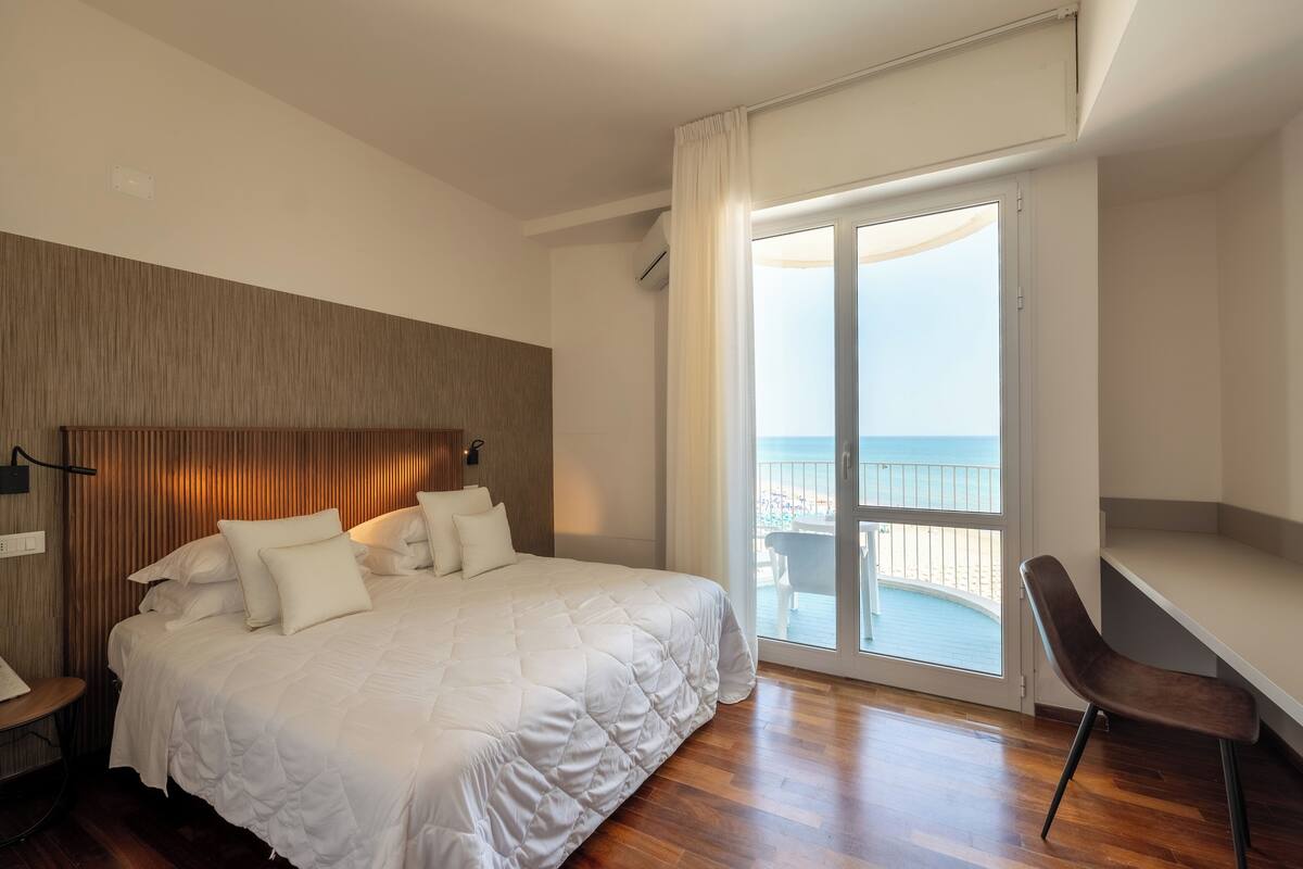 A hotel room with a large bed, a desk, and a balcony with a view of the sea.