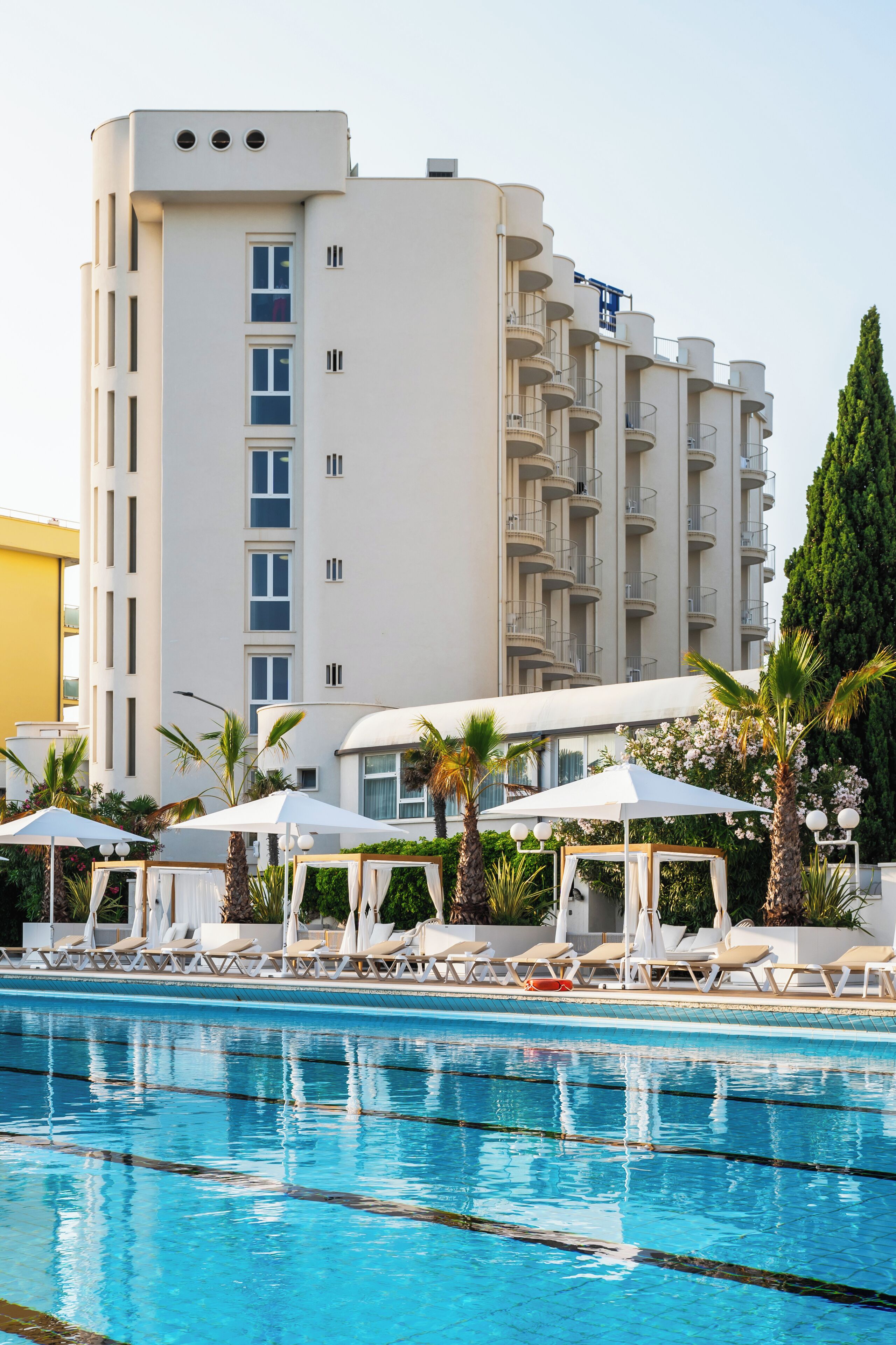 A swimming pool with lounge chairs and umbrellas, surrounded by palm trees and a modern building with balconies.