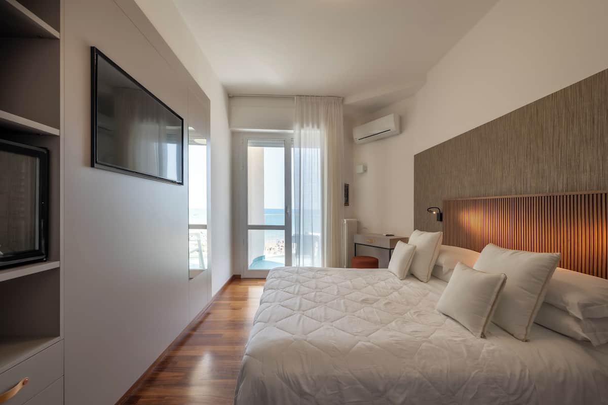 A modern bedroom with a large bed, a flat-screen TV, a wooden headboard, and a view of the balcony.