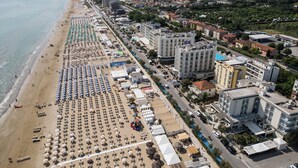 An aerial view of a coastal city with a long beach lined with rows of sunbeds and umbrellas, adjacent to a dense urban area with high-rise buildings and a clear road. - HR Resort (Senigallia)