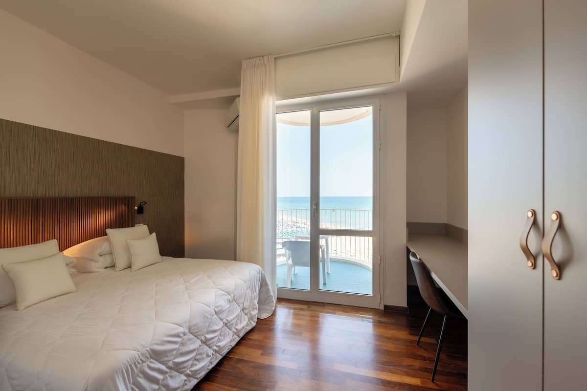 A hotel room with a large bed, a wooden headboard, a balcony with a view of the sea, and a desk with a chair.
