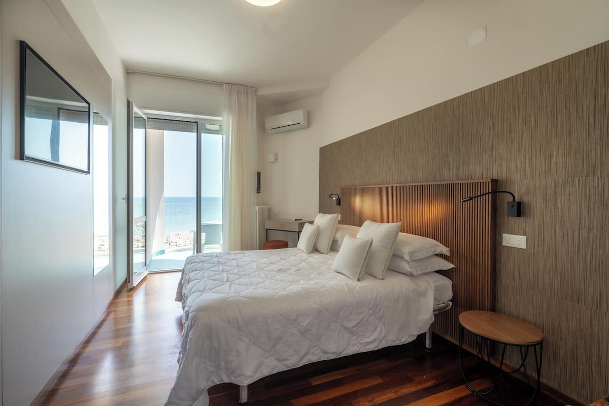 A modern bedroom with a large bed, a wooden headboard, a small round table, and a view of the sea through a sliding door.