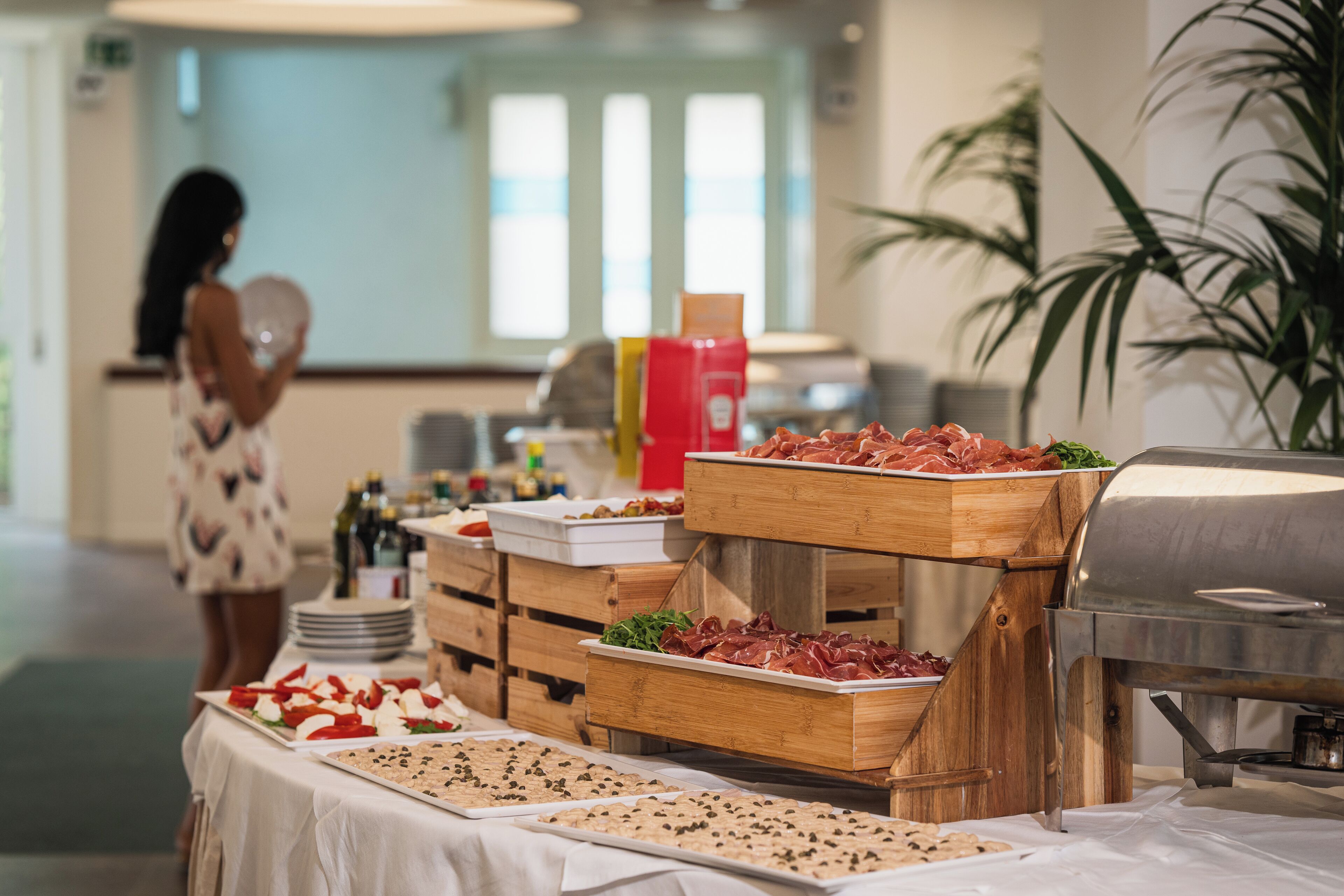 A buffet setup with various food items and a grill, in a room with a large window and a plant.