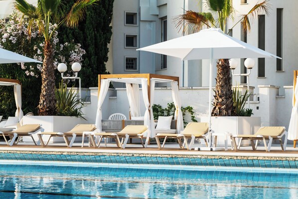 Zona junto a la piscina con tumbonas y sombrillas, una palmera y un edificio al fondo.