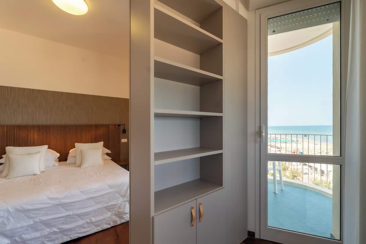 A bedroom with a bed, a built-in shelving unit, and a balcony with a view of the beach.