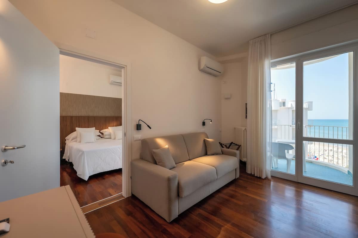 A modern hotel room with a wooden floor, a beige sofa, a bed with white bedding, and a balcony with a view of the beach.