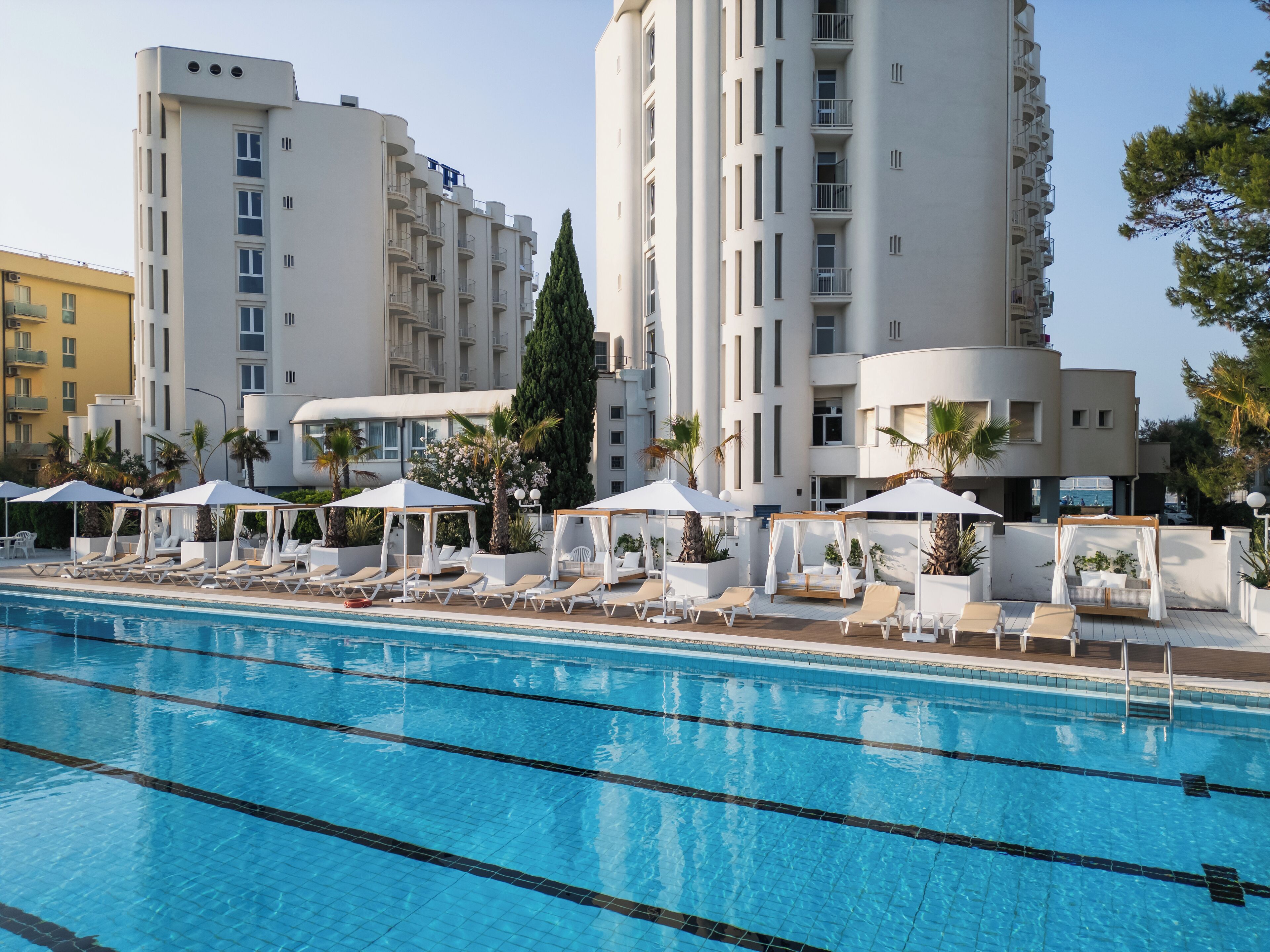 Una piscina con lettini e ombrelloni, circondata da edifici moderni con balconi.