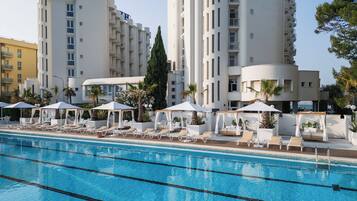 Una piscina con lettini e ombrelloni, circondata da edifici moderni con balconi.