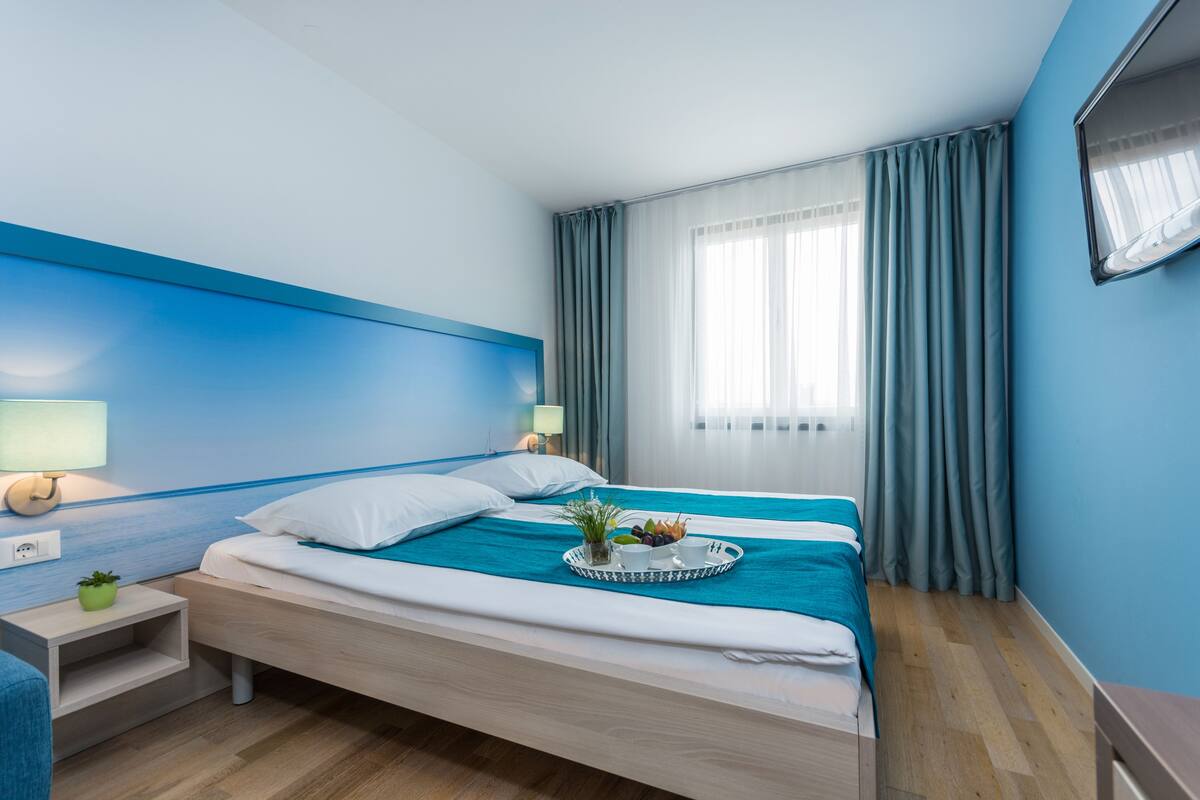 A modern hotel room with a bed, bedside tables, a window with curtains, and a wall-mounted TV.