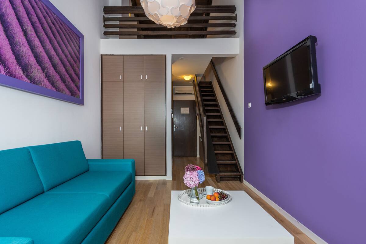 A modern living room with a teal sofa, a white coffee table with a floral arrangement, a large purple wall, a TV mounted on the wall, and a staircase leading to an upper level.