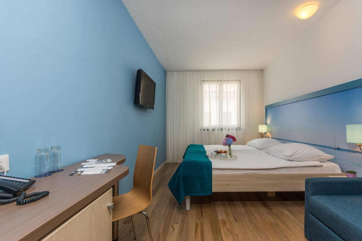 A hotel room with a bed, a desk, a chair, a TV, and a window with curtains.