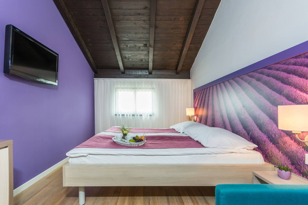 A bedroom with a bed, a flat-screen TV mounted on the wall, a window with sheer curtains, and a wall mural.