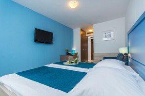 A hotel room with two beds, a flat-screen TV, a desk with a chair, a small table with a tray of fruit, and a picture on the wall. - Hotel Vista (Vrsar)