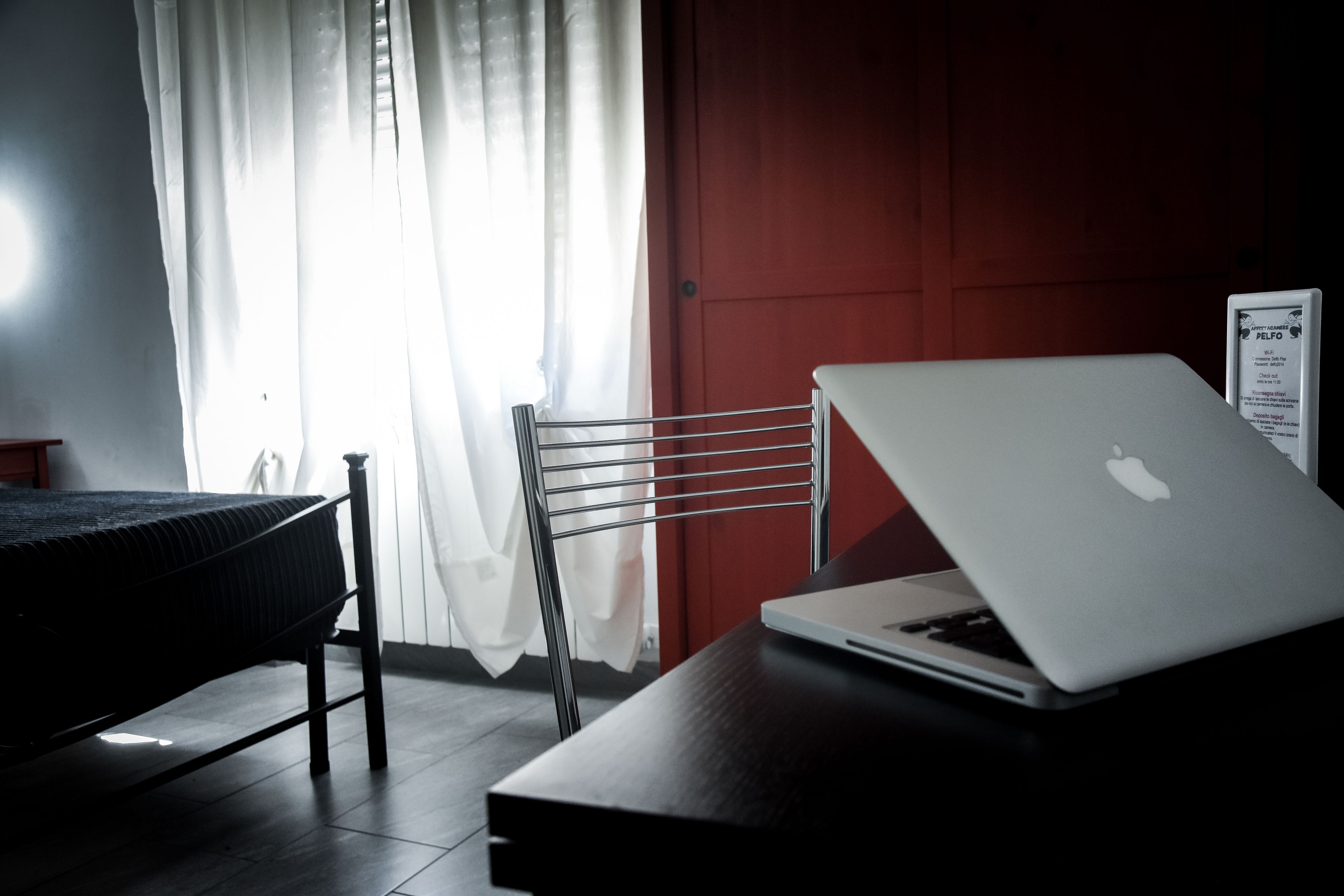 A room with a bed, a radiator, a desk with a laptop, a chair, and a red door.