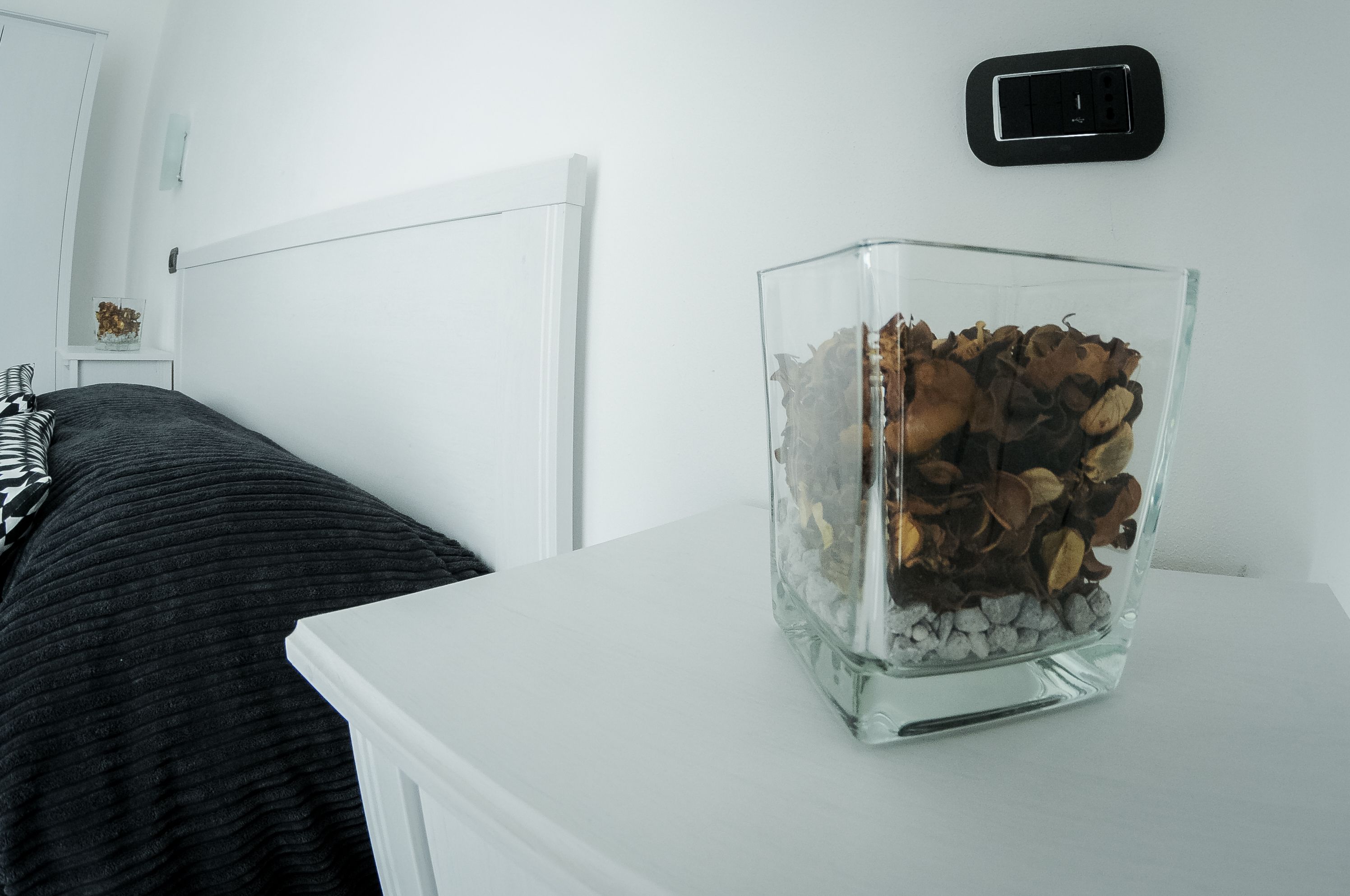 Eine klare Glasvase mit getrockneten Blumen auf einem weißen Nachttisch in einem Raum mit weißen Wänden und einem weißen Kopfteil.