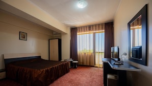 A hotel room with a bed, a desk with a television, a small table, and a window with curtains. - Coral Eforie Nord (Eforie)