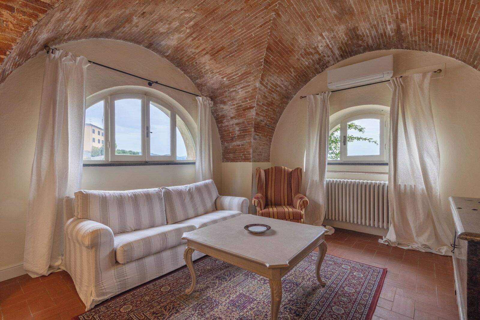 A cozy room with a sofa, armchair, and table, featuring arched windows and a brick ceiling.