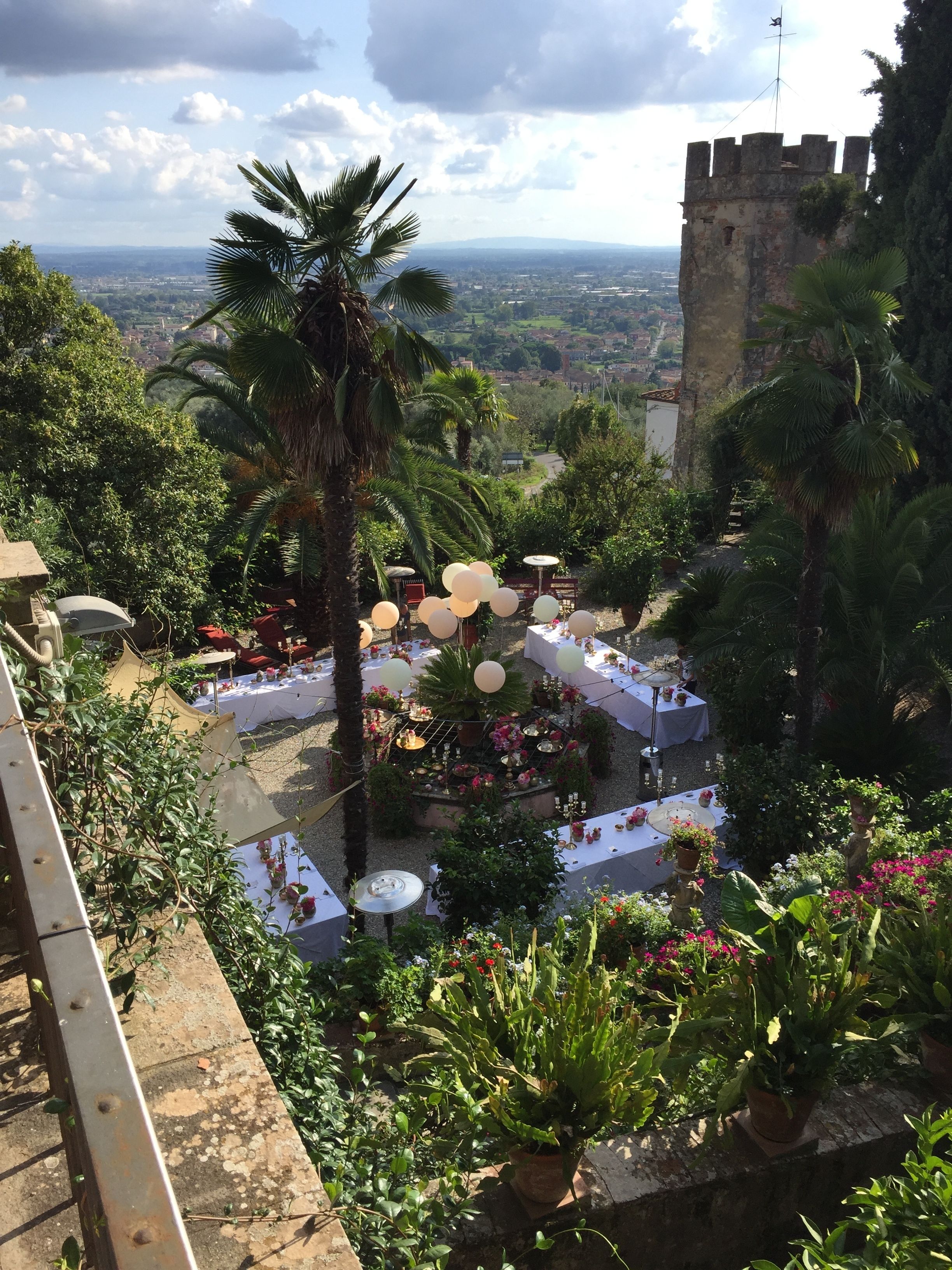 A garden setting with tables and chairs, surrounded by lush greenery and a view of a distant cityscape.