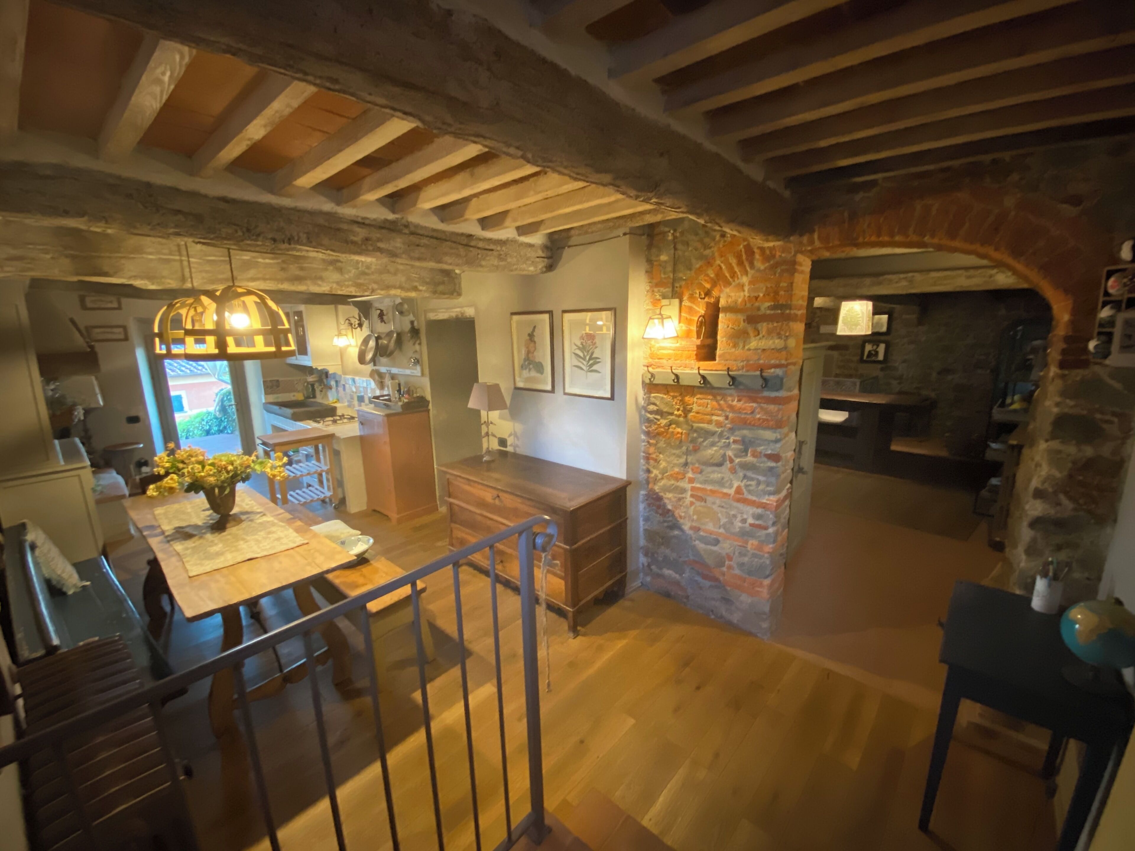 Ein rustikales Interieur mit sichtbaren Holzbalken, einem Steinkamin, Holzdielen und einem Essbereich mit einem Blumenstrauß.