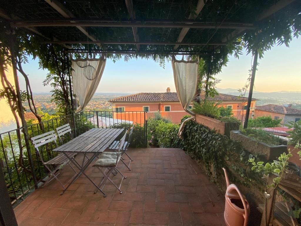A covered patio with a table and chairs, a view of the cityscape, and a pergola with vines.
