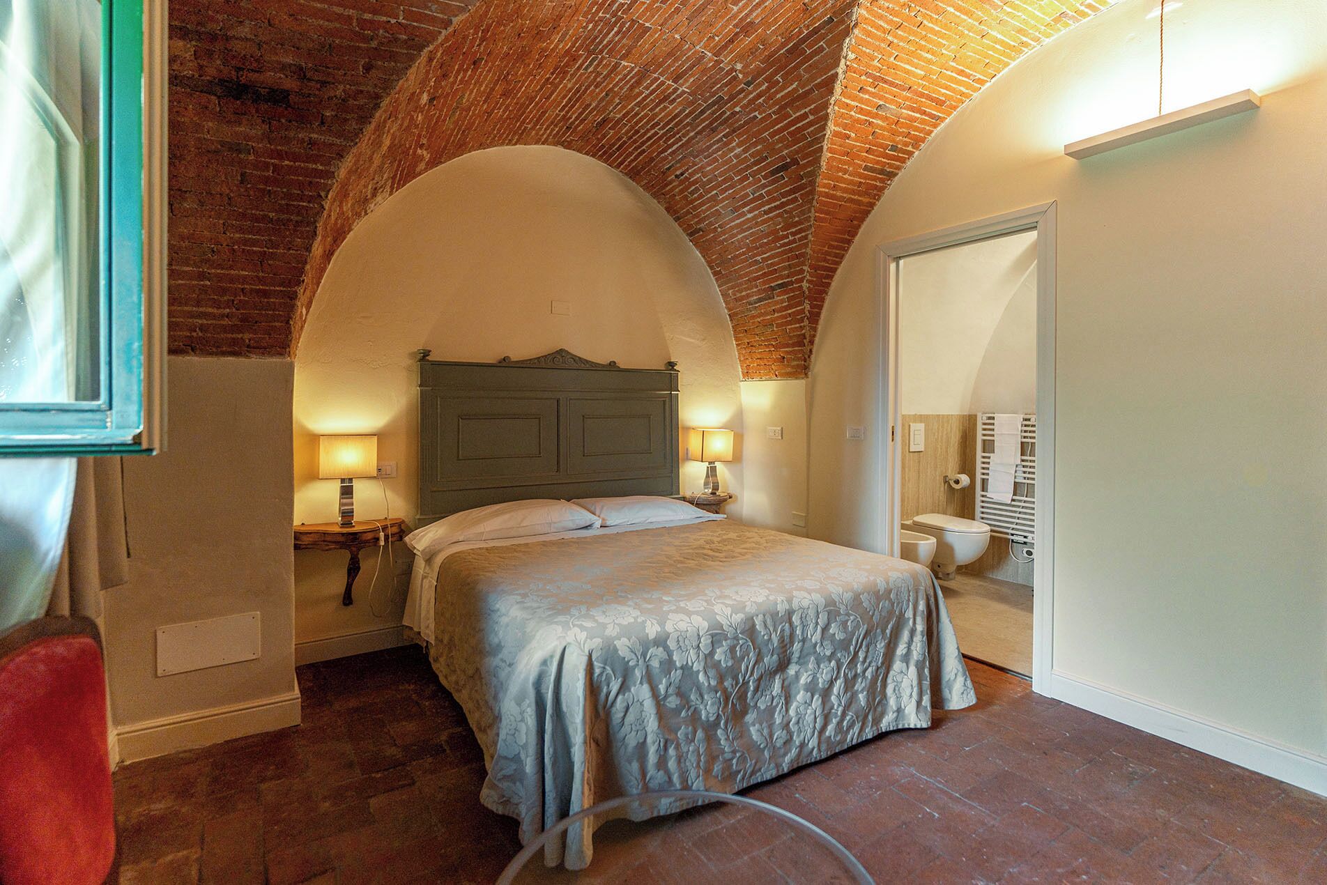 A bedroom with a brick ceiling, a bed with a floral bedspread, two bedside lamps, and a white door.