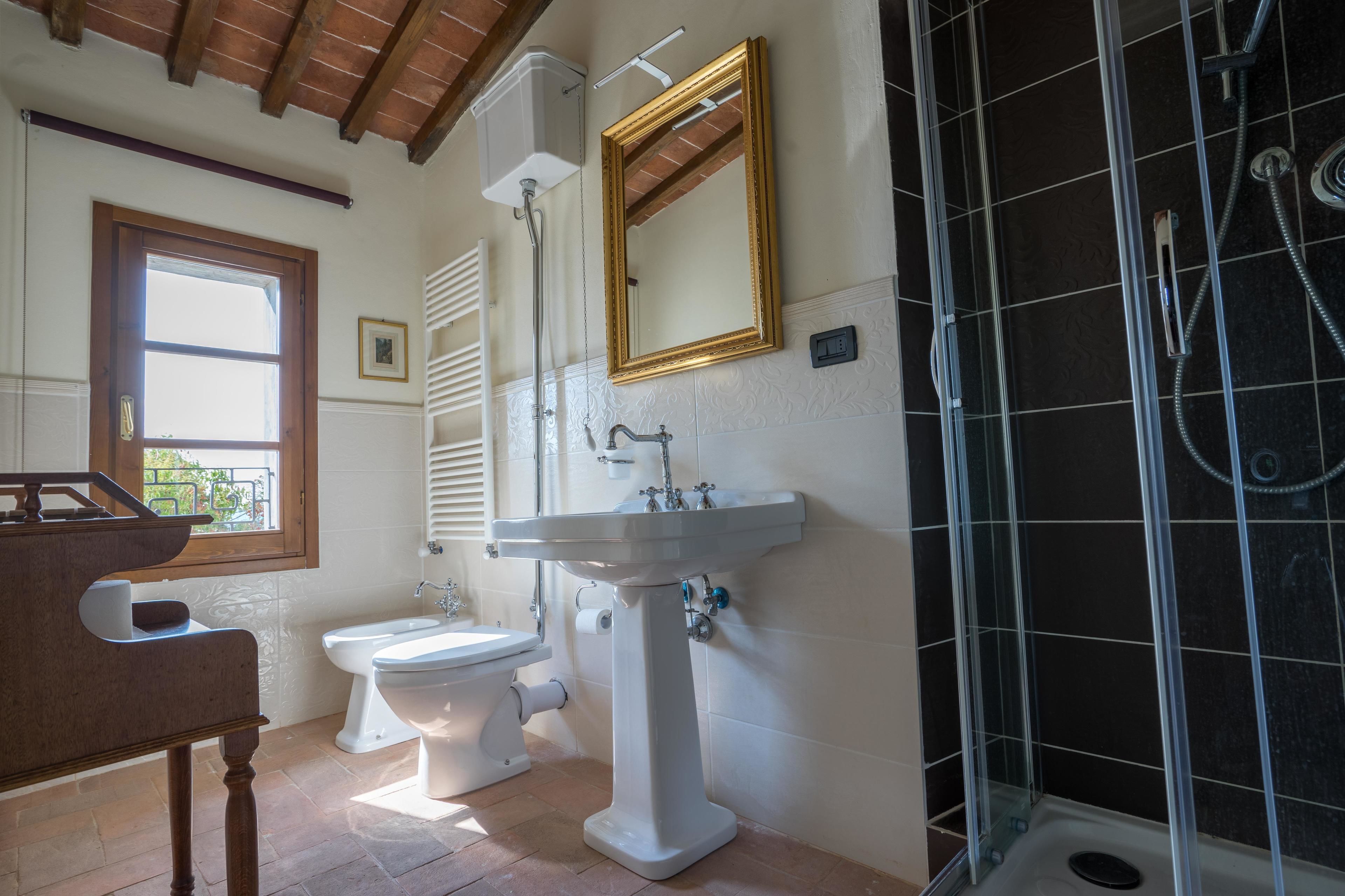 A bathroom with a pedestal sink, toilet, and shower. There is a window with blinds and a wooden ceiling.