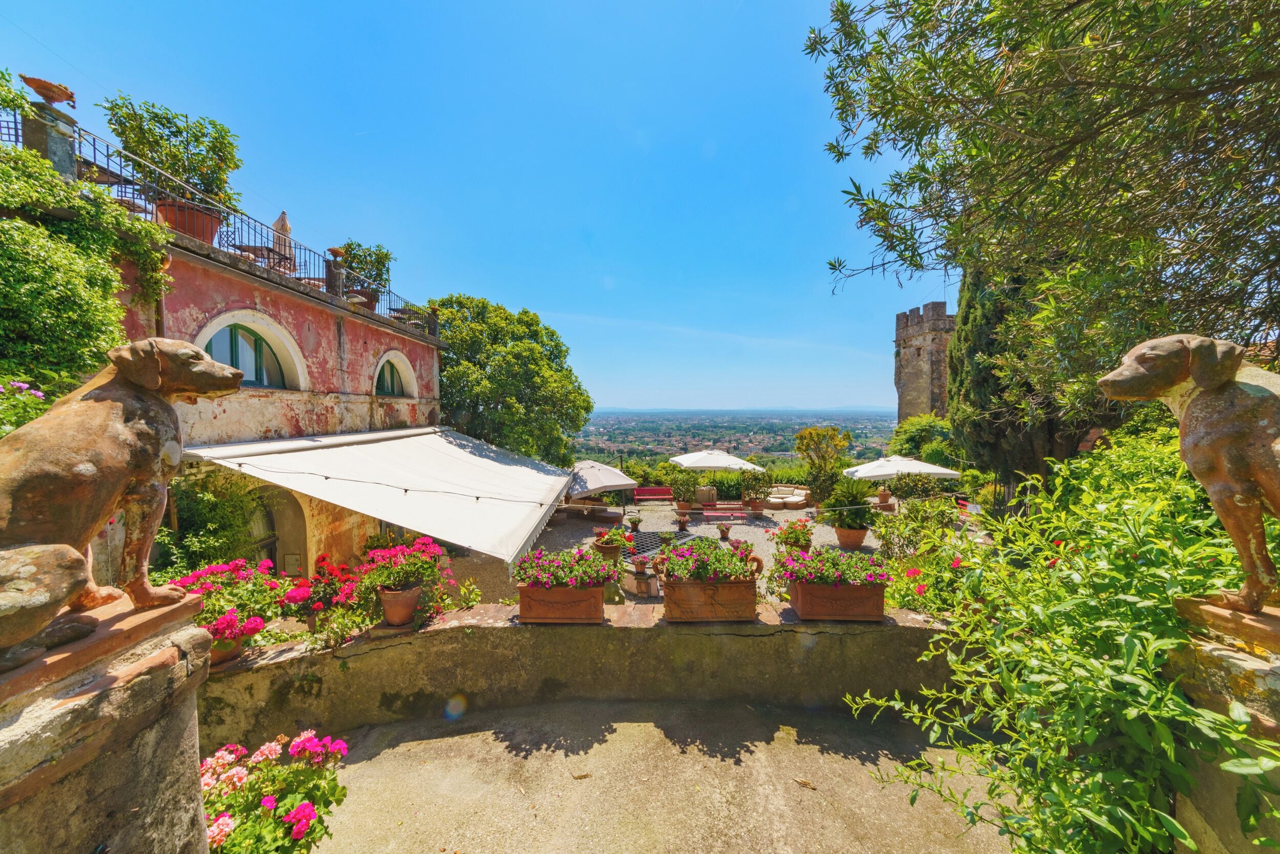 Un patio con estatuas de piedra, plantas en macetas y vistas a un castillo lejano.