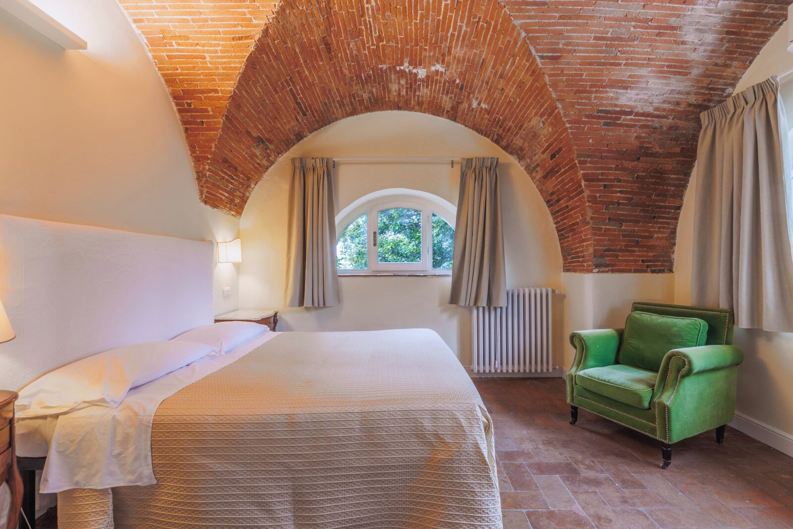 Quarto com teto de tijolos, uma cama grande, poltrona verde e janela com cortinas.