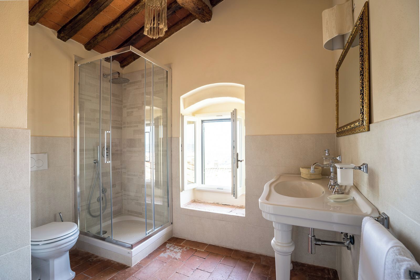 Un bagno con cabina doccia in vetro, un lavandino bianco, un water e una finestra con porta aperta.