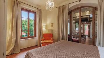 A bedroom with a large bed, a chair, a chandelier, and a view of the outdoors through a window.