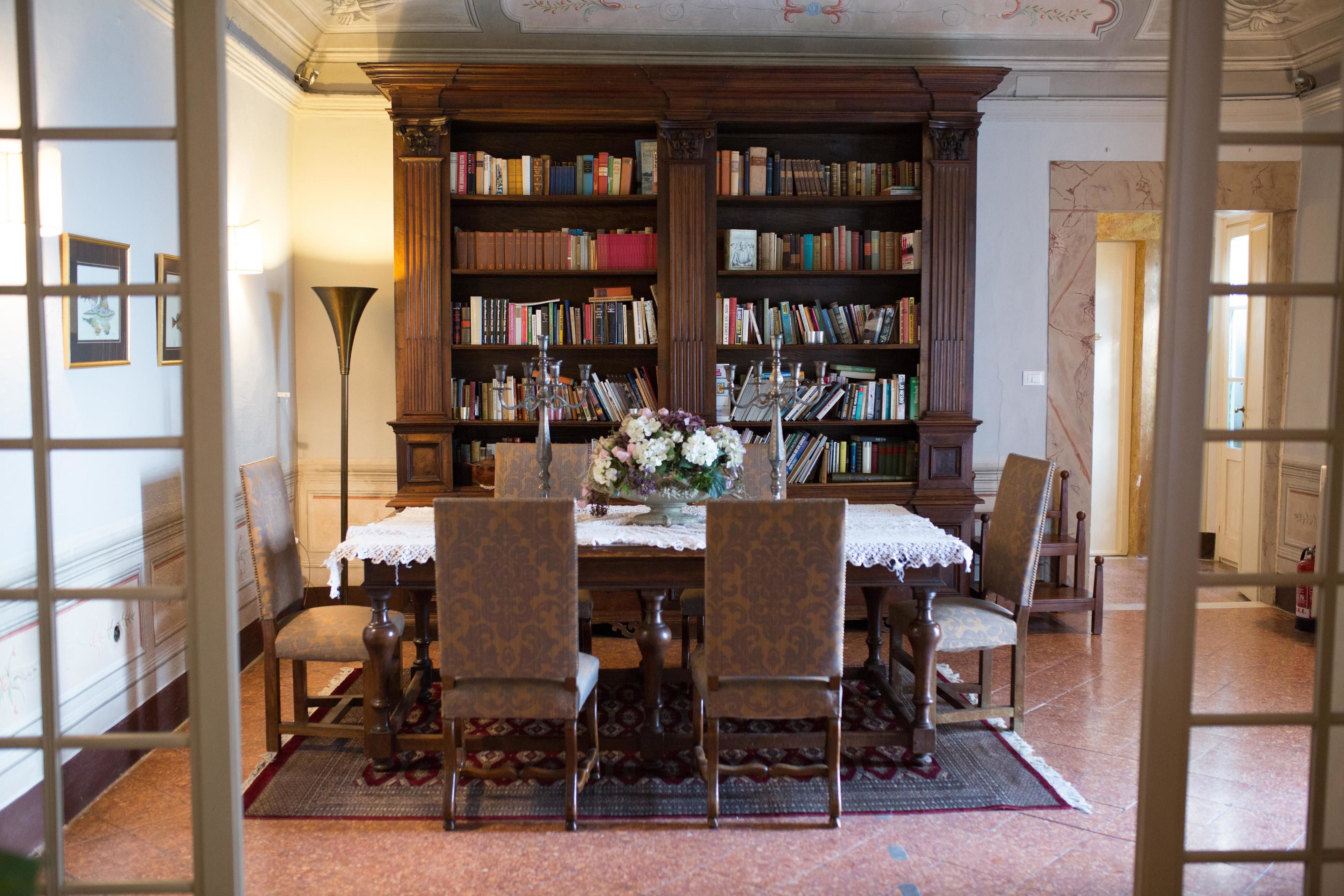 Una biblioteca con una amplia estantería de madera llena de libros, una mesa con mantel de encaje y sillas.