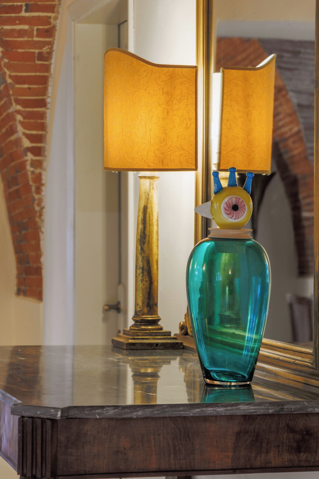 A teal glass vase on a wooden surface with a lamp and mirror in the background.