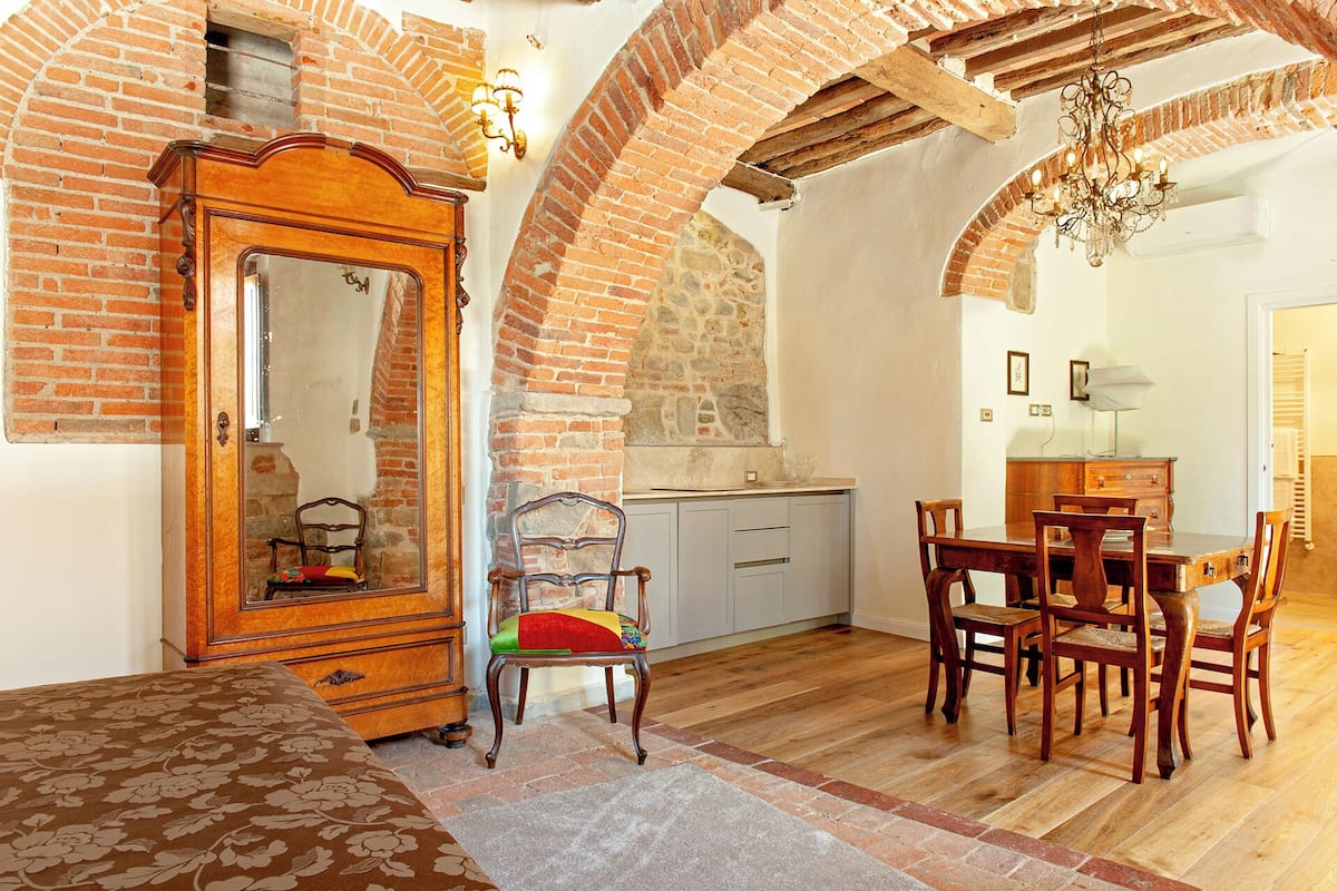 Una habitación con un armario de madera, una mesa de comedor con sillas y una zona de cocina con pared de piedra y arco de ladrillo.