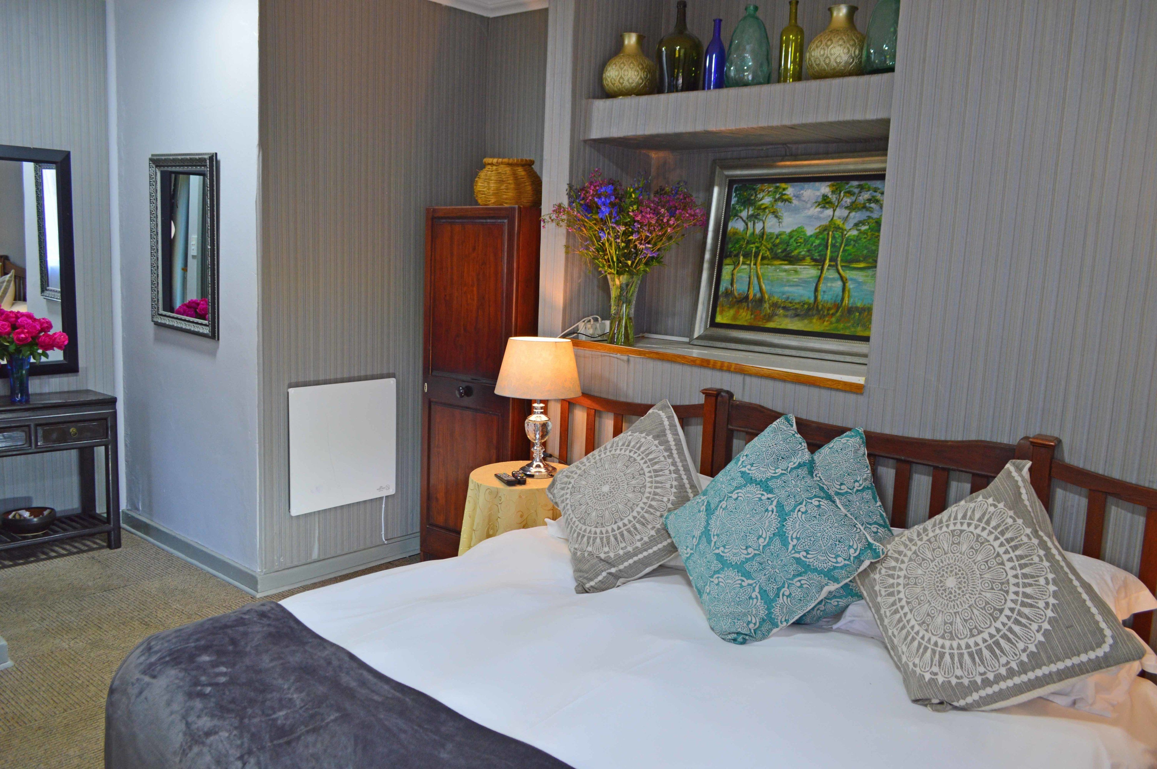Una habitación de hotel ordenada con cama, mesita de noche con lámpara, espejo, cómoda y estante con adornos.