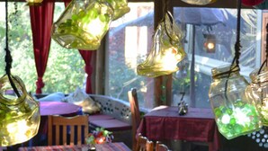 Des bocaux en verre suspendus, contenant une substance verte, une chaise en bois, une table recouverte d’une nappe rouge et une fenêtre donnant sur un paysage verdoyant.