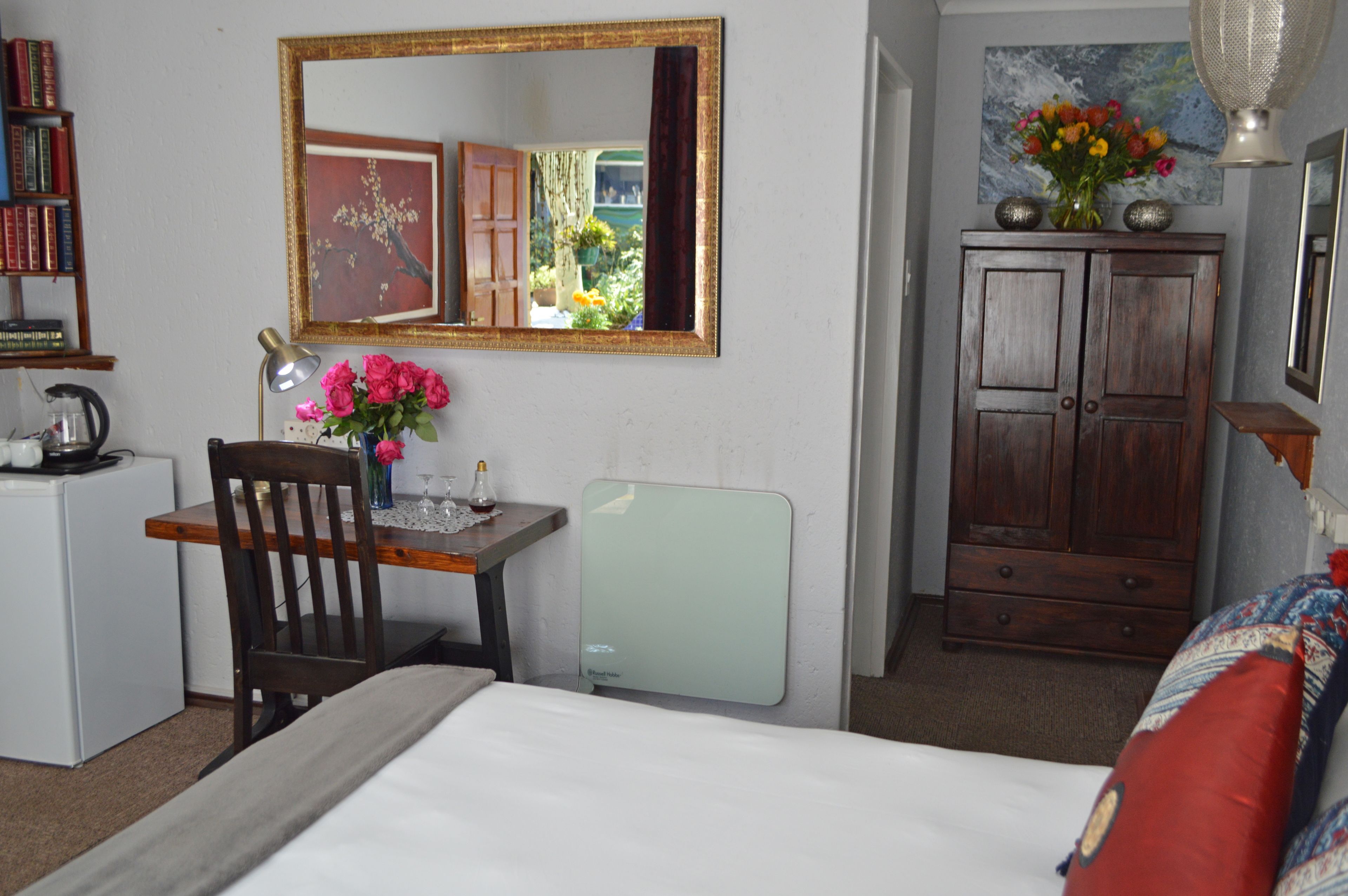 A hotel room with a bed, a desk with a chair, a dresser, a mirror, and a bookshelf.